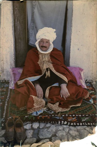 A turbanned man, identified as a Sheik, is sitting on a patterned rug on the ground in front of a doorway. He is wearing a red robe with elaborate gold trim, two tassels and a blue lining. His shoes are placed in front of him on the rug, in the foreground. There is a blue curtain hanging in the doorway.