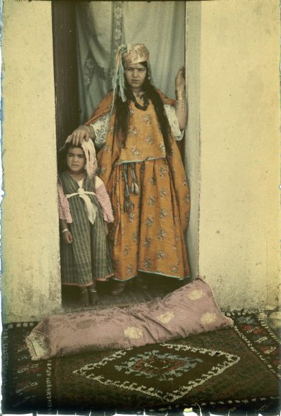A woman is standing in a doorway with her right hand resting on the head of a small girl. Both of them are wearing traditional dress with head scarves. The women has tattoos on her forearms. There is a long rectangular pillow lying on a patterned carpet in front of the doorway, which has a blue curtain hanging inside.