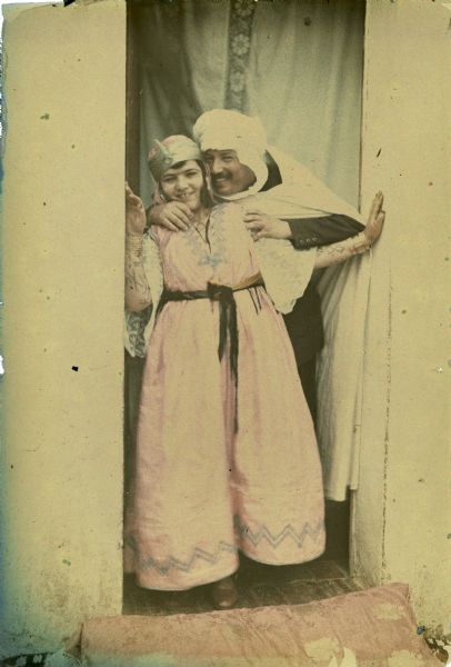 A man, identified only as "Sheik" is posing with his wife in a doorway with a curtain hanging behind them. The man is wearing a white robe over a Western-style suit, and a turban. His wife has multiple bracelets on her tattooed forearms, and is wearing a traditional dress. A pillow is in front of the doorway in the foreground.