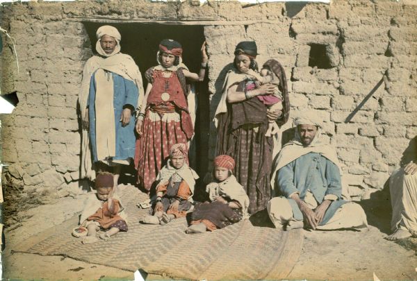A group identified by the photographer as "two couples, four kids" are posing in front of a brick dwelling. One man and woman are standing in the doorway; to the right, a woman is holding an infant in her arms, and the second man is sitting on the ground. Three children are sitting on a carpet on the ground. On the far right is the hand, foot, and leg of another adult who is sitting. The women have facial and forearm tattoos. The men are wearing turbans, capes, and blue coats with buttons. None of the subjects are wearing shoes.