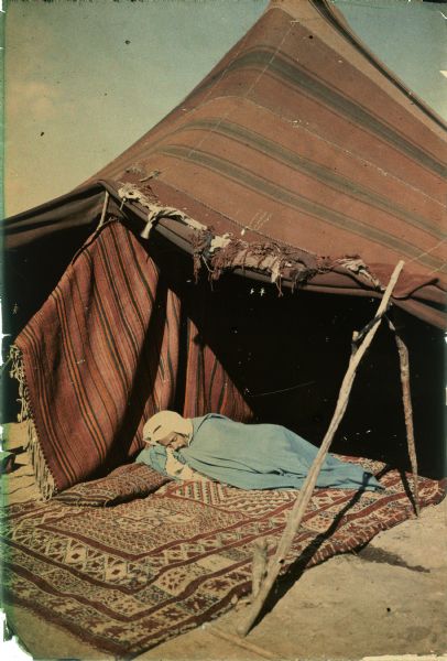A man, wearing a white turban and a blue cape or blanket wrapped around him, is sleeping on pillows and a carpet in front of a tent. A carpet has been draped over one of the tent supports as a screen or wind break.