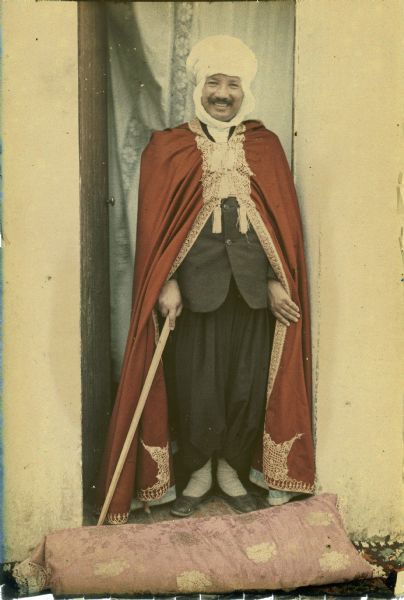 A man wearing a turban and cape with gold trim is standing and smiling in an open doorway, with a curtain hanging behind him and a pillow in front of his feet. He is wearing traditional trousers with a Western style suit jacket, and has a cane in his right hand.