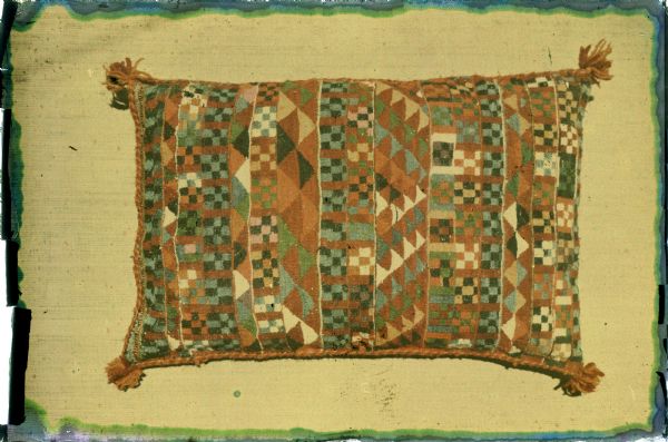 View of a flat weave pillow with geometric patterns and cord detail on the edges, and small tassels at the corners.