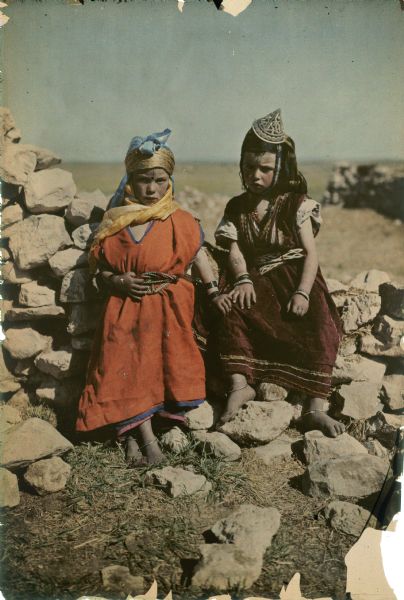 Two young girls are posing outdoors wearing traditional dress. The girl on the left is standing and leaning against the stone wall behind her, and the second girl is sitting on a pile of stones. Both girls have facial and arm tattoos, and are wearing headscarves and wrist and ankle bracelets. The girl on the right is wearing a conical hat.