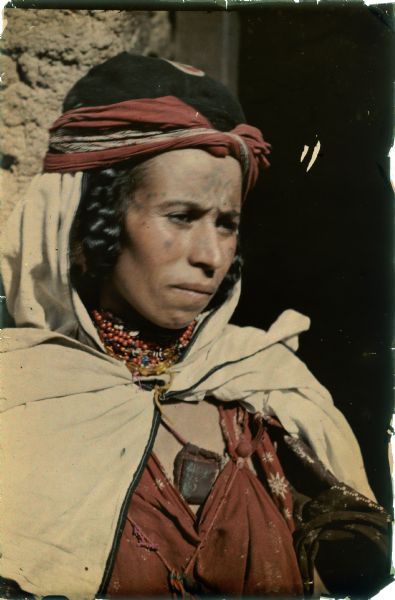 A woman is posing for a head and shoulders portrait in the doorway of a dwelling. She is wearing traditional dress with head scarf and a bead necklace. Her hair is in braids.