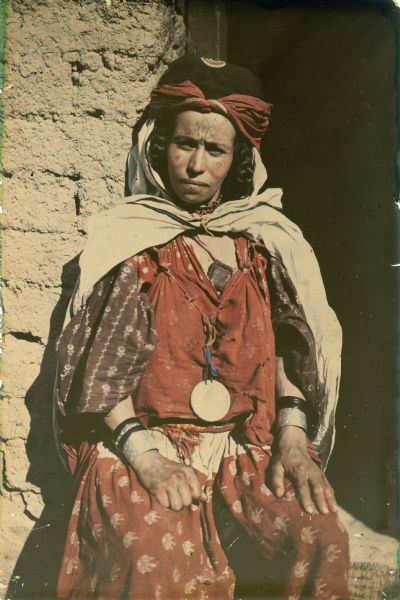 A woman in traditional dress is posing in a doorway for a three-quarters seated portrait. She has facial and forearm tattoos, a beaded necklace and two pendants. Her hair is in braids and she is wearing a headscarf.