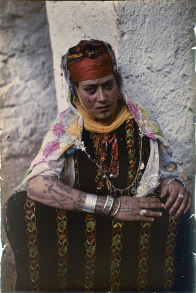Portrait of a woman sitting on the ground against a white plaster wall. She is wearing a richly embroidered dress and shawl, and a headdress with metal bangles. She has facial and forearm tattoos. Her jewelry includes multiple bracelets, a beaded necklace, and two rings.