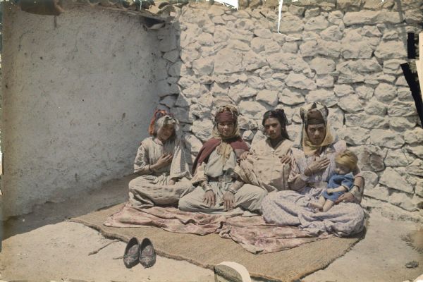 Four young women, one holding an infant, are posing sitting on a rug on the ground, inside the corner of two walls. Identified by the photographer only as "four wives," they are wearing traditional dress, multiple bracelets, and head scarves. Most of the women have facial and forearm tattoos. A pair of shoes have been placed near edge of the the rug.  