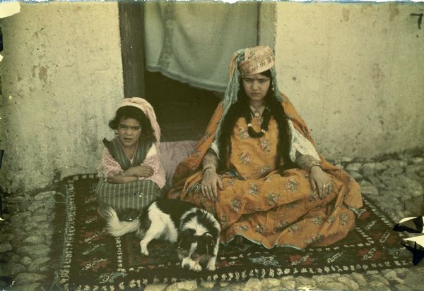 A woman and young girl are posing, sitting on the ground on a patterned carpet, with a dog resting with them. The woman has forearm tattoos, multiple bracelets and rings. The door into an adobe dwelling is behind them.