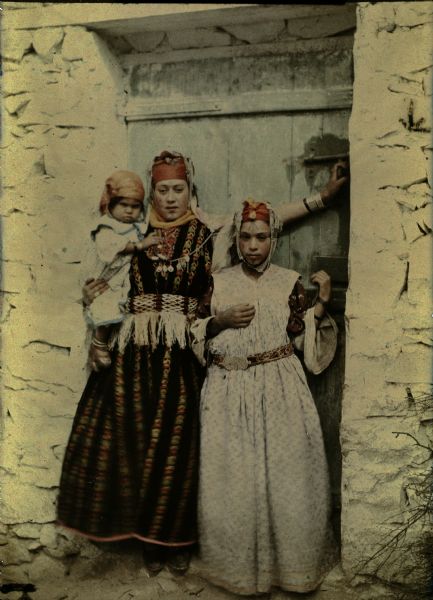 Two women, identified by the photographer as "wives" (possibly both wives of the same man), posing in the doorway of a stone building. The wooden door behind them is painted blue. The woman on the left is holding a young child on her hip. All three of them are wearing headscarves and are in traditional dress. The women have facial tattoos. 