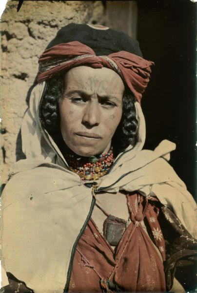 An unidentified woman is posing for a head and shoulders portrait in the doorway of a stone or adobe building. She is wearing a head scarf, beaded necklace and traditional dress. Her hair is in braids and she has facial tattoos.  