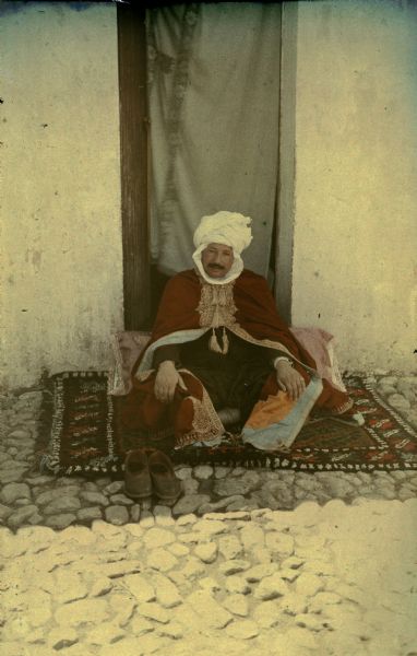 An man, identified only as a sheik, sitting on a patterned carpet in front of a narrow doorway. There is a large pillow behind him. He is wearing a white turban and cloak embroidered in gold. His shoes are sitting on the ground in front of the carpet.