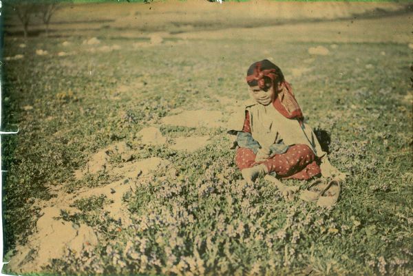 A young girl is smiling as she is posing while sitting in a rocky field of wildflowers with purple blossoms. She is wearing traditional dress and a head scarf. Her shoes are on the ground in front of her.