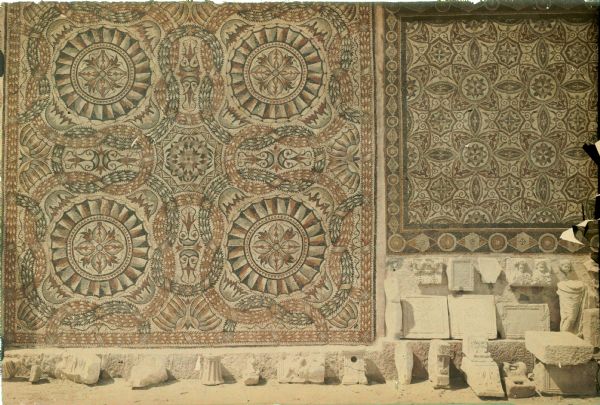 Roman carvings and architectural remnants are displayed out-of-doors below two intricate mosaics in Algeria, possibly in Constantine.