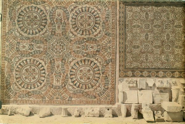 Roman carvings and architectural remnants are displayed out-of-doors below two intricate mosaics in Algeria, possibly in Constantine.