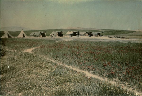 View across field towards an automobile parked in front of a row of tents set up in a landscape of rolling hills. Two men are sitting at a table near the third tent from the left. Poppies and other wild flowers are blooming in profusion in the foreground.