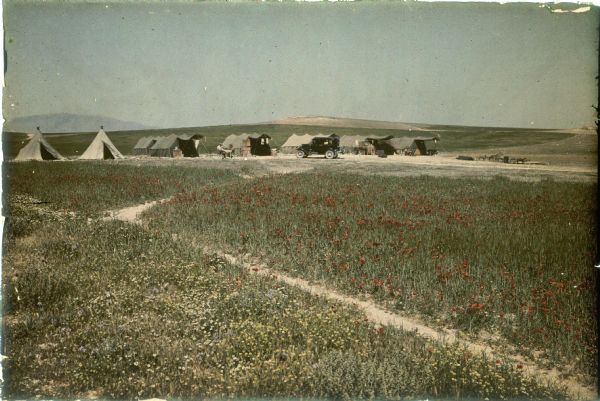 View across field towards an automobile, possibly a 1930 model Citroen, parked in front of a row of tents set up in a landscape of rolling hills. Two men are sitting at a table near the third tent from the left. Poppies and other wild flowers are blooming in profusion in the foreground.