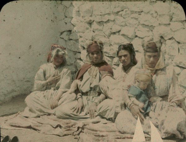 Four young women, one holding an infant, are posing sitting on a rug on the ground, inside the corner of two walls. Identified by the photographer only as "four wives", they are wearing traditional dress, multiple bracelets, and head scarves. Most of the women have facial and forearm tattoos. A pair of shoes have been placed near edge of the the rug at lower left.  