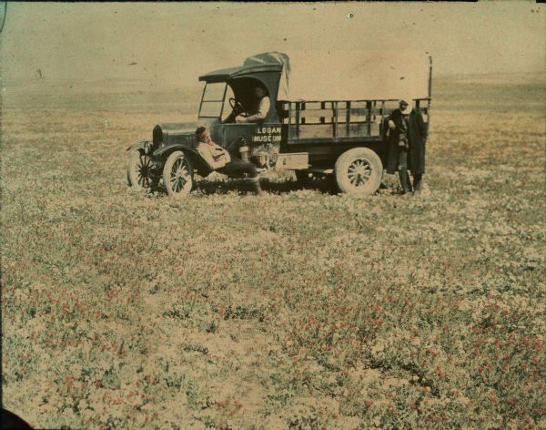 A 1925 Ford Model T Roadster pickup is parked in a field of red-blooming wildflowers. There is a man in the driver's seat, another man is reclining on the fender and running board. A third man, who is wearing a turban, is standing near the rear of the truck with his arm resting on one of the sideboards. A partially obscured sign behind the driver's door reads: "Logan Muséum Beloit College."