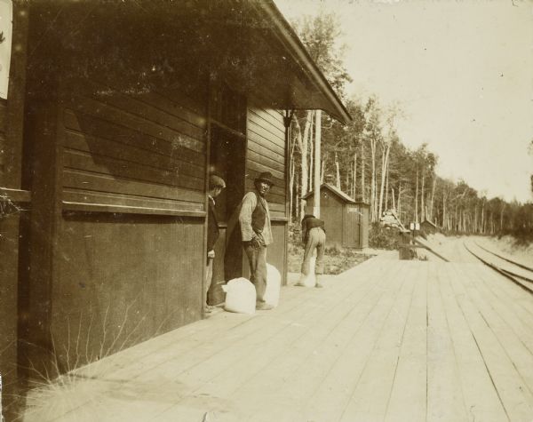 View down platform of Lakewood train station, with two men standing near a doorway on the left near two sacks. Another man is further down the platform bending over another sack. A smaller building in the background has a sign on the door for "Men." There are railroad tracks on the right.