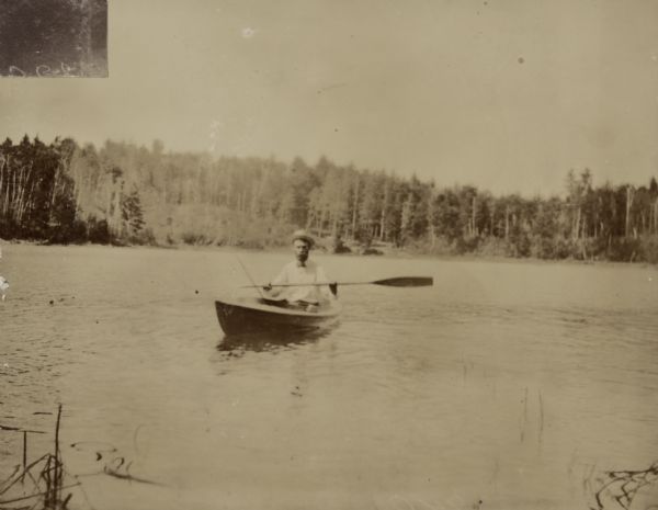View across the water towards George Holt paddling a clinker-built duck boat on Archibald Lake. Caption reads: "At sea."