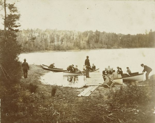 A group of Holt family members and guests is arriving at the Island on Archibald Lake, with lumberjacks from Holt Lumber Company rowing the boats. The man standing on the pier is holding an instrument case. Caption reads: "Arrival at Island Lodge, Aug 24, 1898." Names of family and guests listed below the photograph, not in order, are: Annie McClure, Nathan Swift, Arthur Blackler, I.P. Rumsey, James McClure, Bess Swift, Bertha Durand, Mary Jackson, Dr. James McClure, Edith Platt, and Wallace Rumsey.