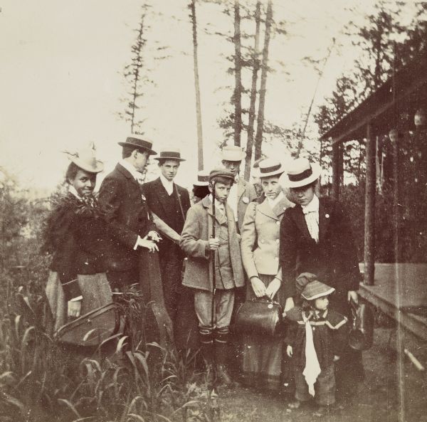 Group portrait of adults and two children standing next to Island Lodge, just before heading home. In the center is Jim McClure, holding a fishing pole. On the right are: Lucy Holt and her daughter Jeannette Holt. This is the first photo of Island Lodge in the collection. Caption reads: "We are homeward bound" and "Jim McClure."