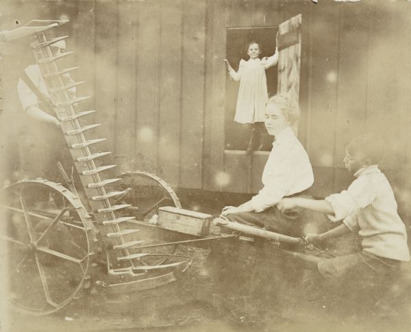 Annie McClure is sitting on the hay mower, with two boys nearby. In the background, Nellie Clapp is standing in the doorway of the barn. The group is at the Farm on the mainland. Caption reads: "Our lady farmers."