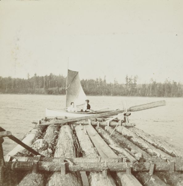 A woman and a boy are sailing the boat named <i>Islander</i> on Archibald Lake. There is a raft in the foreground. Caption reads: "An Ingenious Affair." 