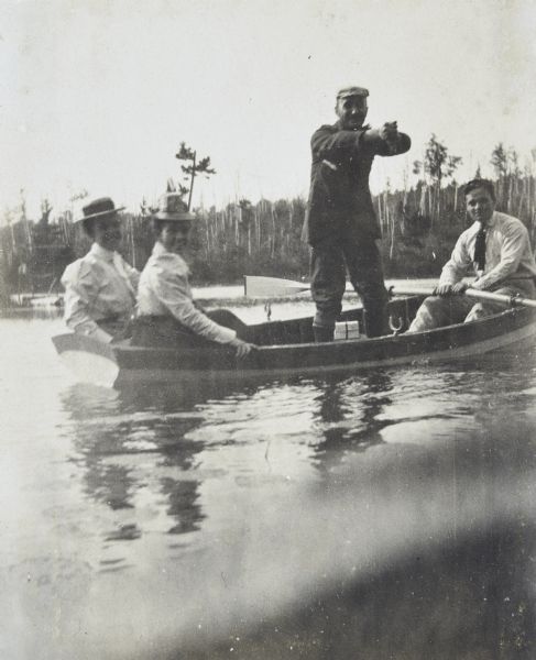 A foursome is boating on Archibald Lake. William Leonard Burnap, standing up in the boat, is wringing water out of something with his hands. The man sitting sitting in the stern of the rowboat is possibly Clint DeWitt. Two women, who are wearing hats, are sitting in the back of the boat. Caption reads: "Squeezing the Tears."