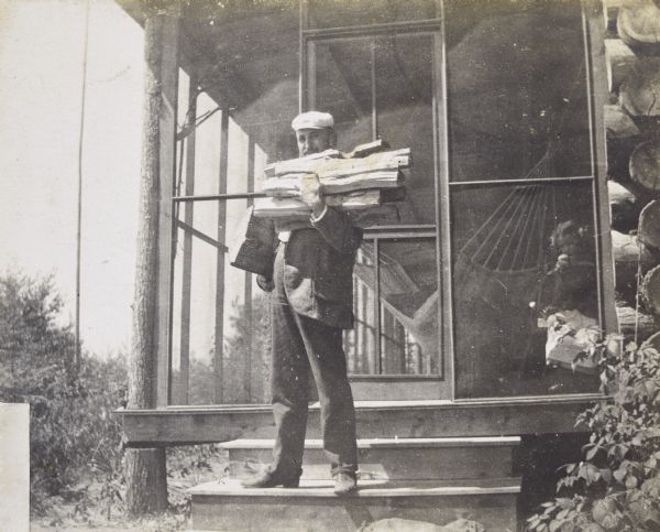 Standing on the steps of Island Lodge, William Leonard Burnap is carrying newly split wood. Jeannette Holt is standing inside the screened-in porch on the right near a hammock. Caption reads: "Earning my board — Mr. Burnap."