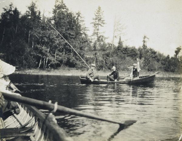 Two clergymen and one boy are sitting in a clinker-built duck boat on Archibald Lake. The clergyman on the left is holding a long cane pole. The clergyman on the right is holding up a fish. Photographer Lucy Rumsey Holt (out of frame) is sitting in a rowboat with an oar in the oarlock in the left foreground. Caption reads: "Two of the clergy... fishing."