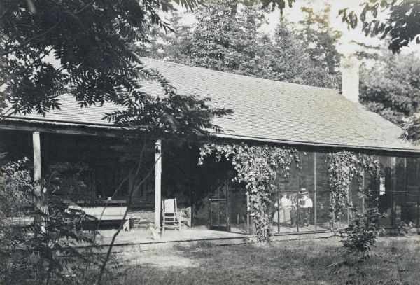 Slightly elevated view of Island Lodge, with a front porch on the left, and a screened-in porch on the right. Lucy Rumsey Holt and Arthur Dana Wheeler are sitting inside. Trees are in the background. Caption reads: "The Lodge, 1907." The caption to a similar photo in Album 6 reads: "Island Lodge. 1907. L.R.H, A.D.W. By Mr. Thiers."