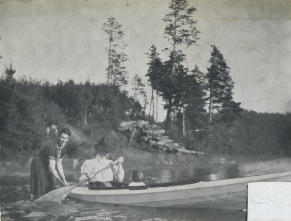 Mother Lucy Holt is paddling the canoe on Archibald Lake with baby daughter Eleanor Holt on board. Another woman, probably a household employee, is standing in knee-deep water holding onto the back of the canoe. In the background, logs are piled on the tree-lined shoreline. Caption reads: "A Safe Swim for Baby Eleanor." 