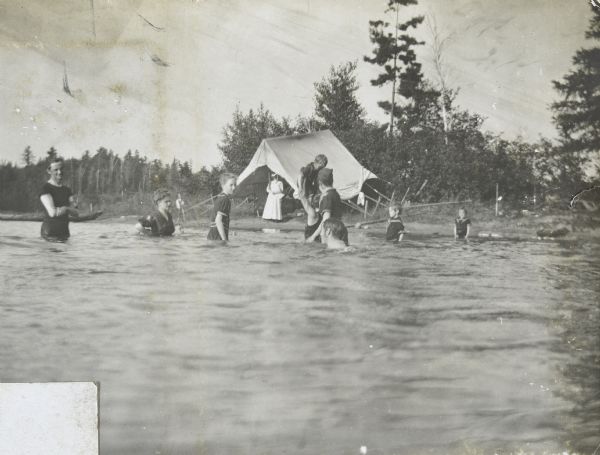 A group of children, supervised by adults, is playing at the bathing beach on Archibald Lake. On the shore is a tent with a woman sitting inside. Caption reads: "A daily frolic." 