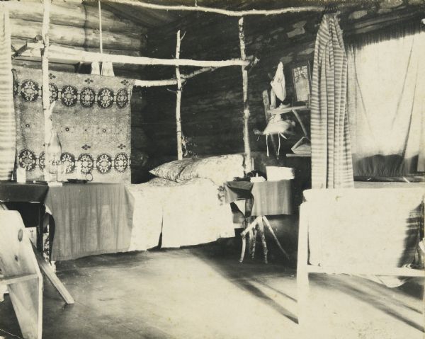 An interior view of the Hermit Cottage, which has a four poster rustic birch bed, bedside table, and a decorative rug hanging from the rear birch post. There is a curtain pushed to the side near the window on the right, which is attached to rings on a birch post that is hanging from the ceiling horizontally in front of the bed. Caption reads: "Favorite Nooks."


