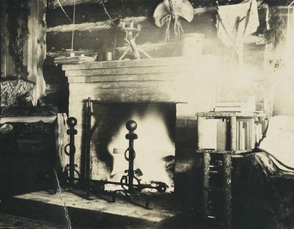 View of the big fireplace ablaze at Island Lodge on Archibald Lake, with screen and andirons. There are various handmade wooden objects on the mantel. On the right side of the fireplace is a bookshelf with books, and a chair. Firewood is stacked on the left side of the fireplace. Caption reads: "Island Lodge."