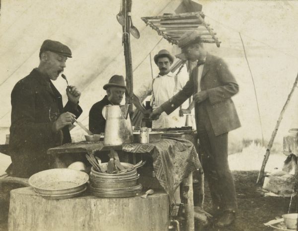 Under a tent, three men are eating a meal prepared by cook Bob Morris, who is standing beside the table. In the foreground is a tree stump which is loaded with tableware and cooking utensils. Caption reads: "A good cook — several good appetites." Names from left to right: "J.F. Rumsey, I.P. Rumsey, Bob Morris — cook, W.A. Holt." 
