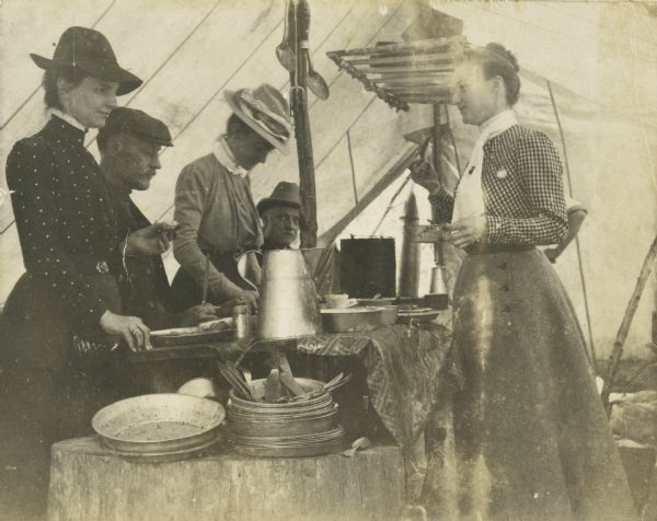 A group of men and women are eating a meal under a tent. Tableware and utensils are on the tree stump in the foreground. Names from left to right: Lucy Rumsey Holt, Joseph Frank Rumsey, Ellen Holt, IP Rumsey, and Minnie May Rumsey. Caption reads: "A good cook — several good appetites. LRH, JFR, Ellen Holt, IPR, MM Rumsey."