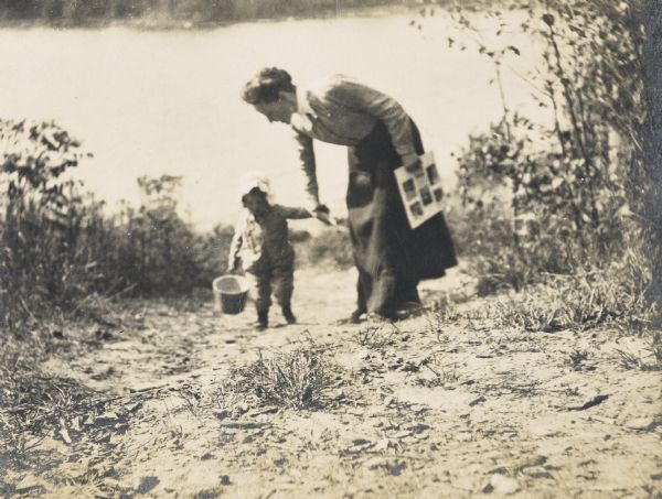Toddler Eleanor Holt, carrying a pail, and Minnie May Rumsey are walking on the path away from Archibald Lake. 