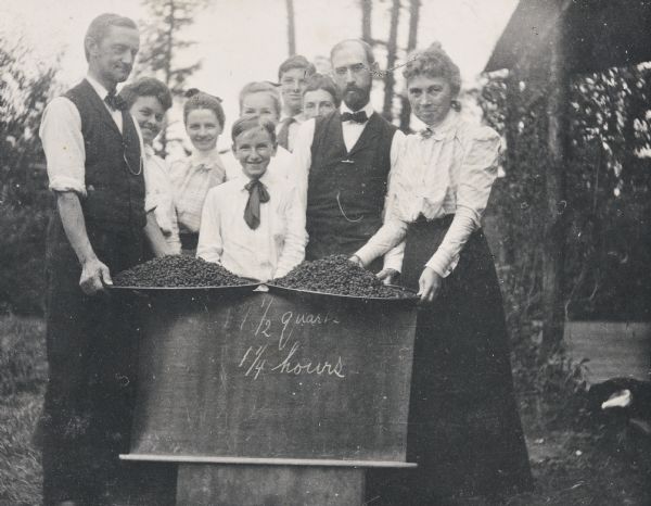 Group portrait of women, men, and children standing in front of a display of freshly-picked blackberries. Dr. James McClure (left) and Miss Smith (right) are each holding a tray of berries. Annie McClure is standing to the right of her father Dr. McClure. Grant Stroh is standing to the left of Miss Smith. A handwritten sign under the trays of berries reads: "17 1/2 quarts, 1 1/4 hours." Photo album page heading reads: "Blackberrying." Names below the photograph are: "Dr. McClure and Miss Smith."
