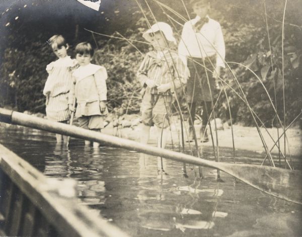 View from rowboat of siblings Jeannette and Alfred Holt standing in the water and watching the rowboat approaching the shore of Archibald Lake. Alfred is holding a pole. Brothers Nathan and Archibald McClure are standing on the right. Names from left to right are: "Jeannette, Alfred, Nathan, and Arch, Sandy Beach."
