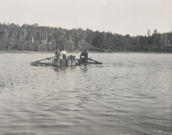 View across water of four men on the baggage raft traveling toward the Island from Apple Tree Landing. Two men, possibly lumberjacks, are rowing. The baggage includes wooden boxes and suitcases. Caption reads: "The baggage raft."


