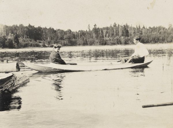 Helen Pool and James McClure are sitting in a canoe on Archibald Lake. McClure is holding a paddle. On the left is the pier. In the foreground, on the right is the oar of a docked boat we can’t see. Names written below the photograph are, from left to right: "Helen, James."

