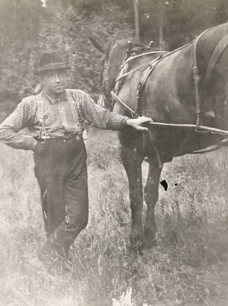 Holding a leather harness, Gus is standing next to a horse in a field. Caption reads: "Gus."
