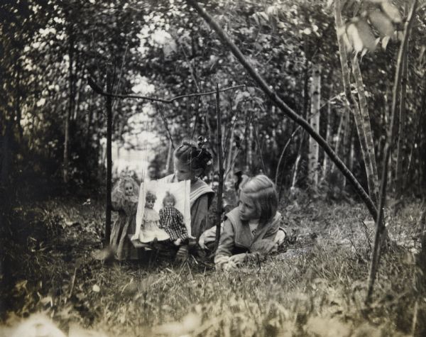 Jeannette Holt and Harriet Stroh are sitting and lying in the grass while playing with three dolls. Two of the dolls are sitting on a swing, made of sticks and hanging from a branch, made of a wood board and fabric. The third doll, standing behind other two, appears ready to push the swing. Archibald Lake is in the background. Caption reads: "Little mothers." 