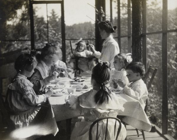 Holt and Stroh children are eating a meal together at a table on the porch at Island Lodge. Names on the right side of the table from front to back: Alfred Holt, Eleanor Holt, Ella, Donald Holt. Ella, a household employee, is feeding Donald Holt in a high chair. Jeannette Holt, with her back to the camera, is sitting at the head of the table. The Stroh girls are sitting on the left side of the table. Caption reads: "Everybody happy. Little Strohs, Little Holts, and Ella. Three times a day."