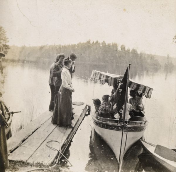 View from shoreline towards a group of children and adults sitting in the <i>Islander</i> steam launch at the Island Lodge pier on Archibald Lake. The Stroh girls are in the boat on the left. Grant Stroh, in profile, is on the right. The <i>Islander</i> flag is flying from the prow of the boat. Three young men and a woman are standing on the pier next to the boat, and there are boats at the shoreline on the left and right. The far shoreline is in the background.
