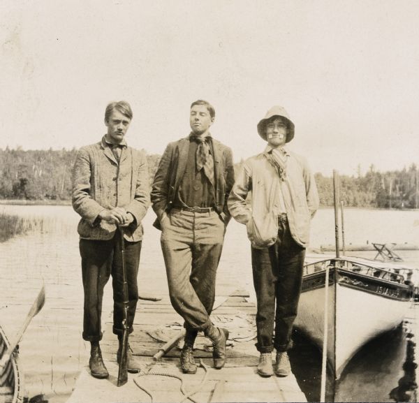Three young men are standing on the pier at Island Lodge on Archibald Lake. The <i>Islander</i> steam launch is tied to the pier with a rope. Holding a gun, Nathan McClure is standing on the left . Wearing a hat and glasses, the young man on the right appears to have tape on his face. A rowboat oar, a rope, and a tarp are lying on the pier. In the foreground on the left is a rowboat.