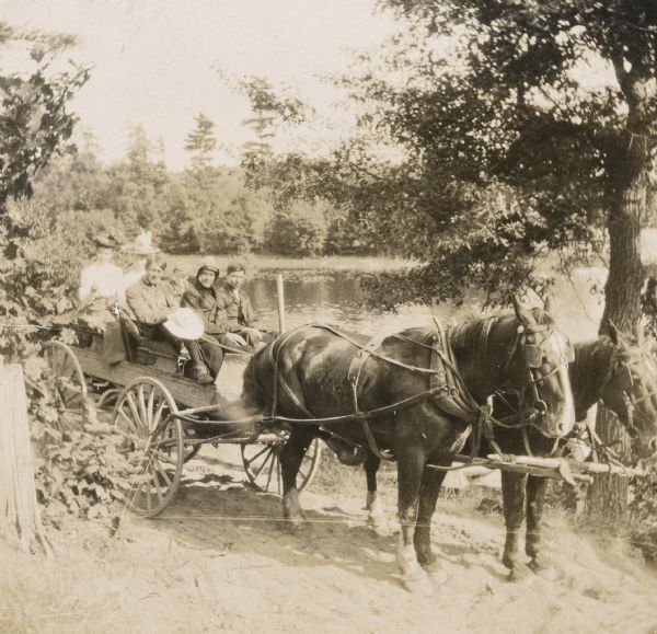 A group of six are riding in a wagon drawn by two horses on a dirt path along the shore of Barn Pond. Nathan McClure is sitting in the front seat on the left side. James McClure is sitting in the front seat on the right side. Two women, including Annie McClure, and a boy are sitting in the back seat. 